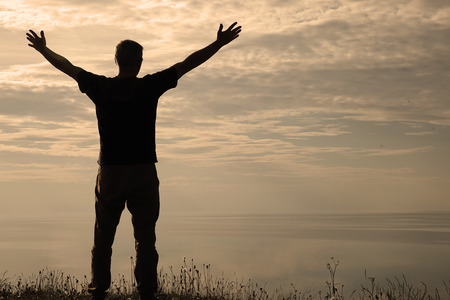 A man stands on top of a mountain and sees the view of nature. The tourist raises his hands and enjoys the fresh air. A man enjoying free happiness in a beautiful landscapeの写真素材