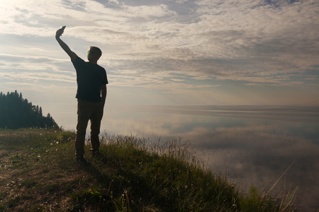 A man standing on top of a hill and looking at his mobile phone in his hands.Silhouette of the traveler.の写真素材