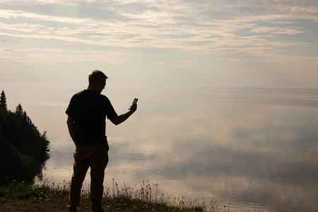 A man standing on top of a hill and looking at his mobile phone in his hands.Silhouette of the traveler. Self.の写真素材