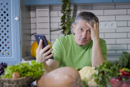 A man in the kitchen prepares vegetarian dishes. Looks up a recipe on the tablet display.の写真素材