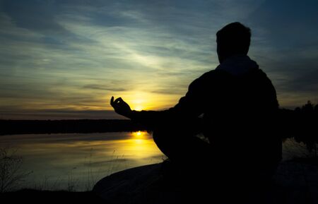 Silhouette of a meditating man . The guy sits on a stone in the Lotus position and looks at the setting sunの写真素材