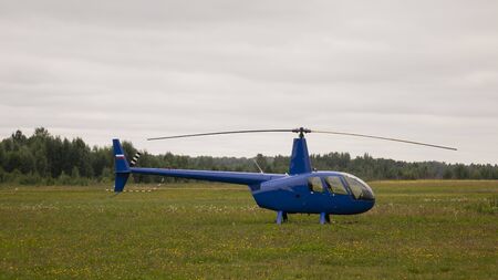 Blue private helicopter on the grass against a cloudy sky.