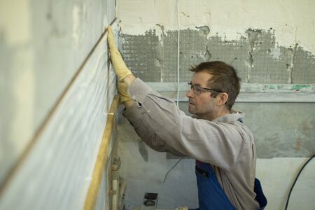 A man in overalls puts a tile in the kitchen. Laying ceramic tiles on grey cement on the wall. The...