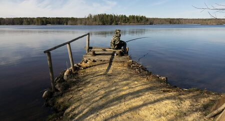 Fisherman in camouflage on the shore of the lake. Fishing with a float rod on the water. A teenager sitting on the riverbank and patiently fishing.の写真素材