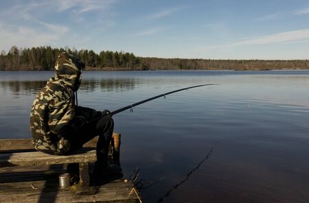 Fisherman in camouflage on the shore of the lake. Fishing with a float rod on the water. A teenager sitting on the riverbank and patiently fishing.の写真素材