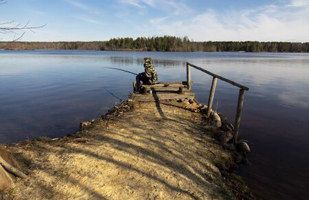 Fisherman in camouflage on the shore of the lake. Fishing with a float rod on the water. A teenager sitting on the riverbank and patiently fishing.の写真素材