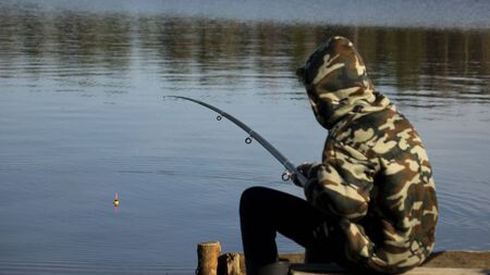Fisherman in camouflage on the shore of the lake. Fishing with a float rod on the water. A teenager sitting on the riverbank and patiently fishing.の写真素材