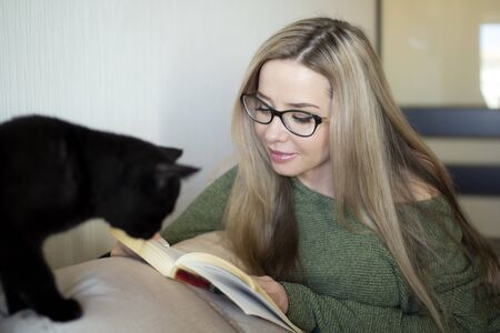 A pretty young woman is happy to read a book at home. The black kitten watches curiously.の写真素材