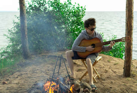 A teenager plays a guitar while sitting on the shore of a lake.の写真素材