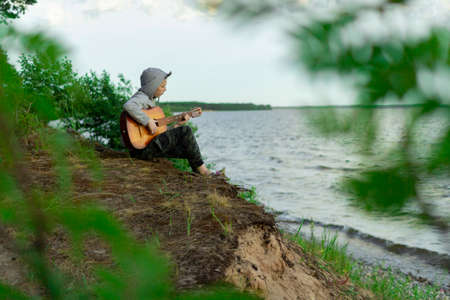 A teenager plays a guitar while sitting on the shore of a lake.の写真素材