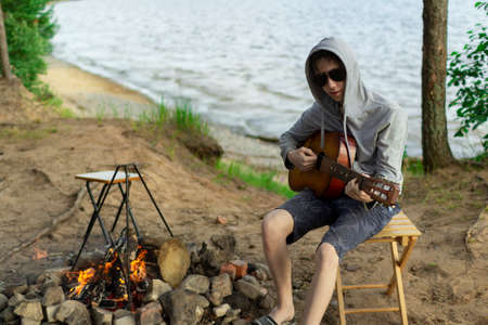 A teenager plays a guitar while sitting on the shore of a lake.の写真素材