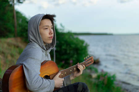 A teenager plays a guitar while sitting on the shore of a lake.の写真素材
