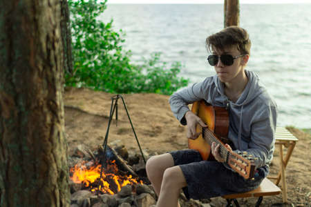 A teenager plays a guitar while sitting on the shore of a lake.の写真素材