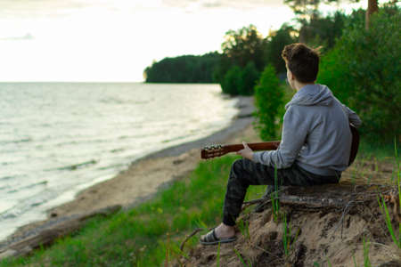 A teenager plays a guitar while sitting on the shore of a lake.の写真素材