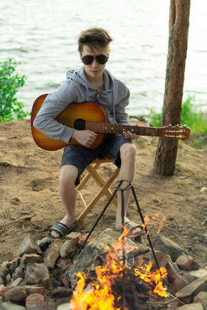 A teenager plays a guitar while sitting on the shore of a lake.の写真素材