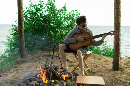A teenager plays a guitar while sitting on the shore of a lake.の写真素材