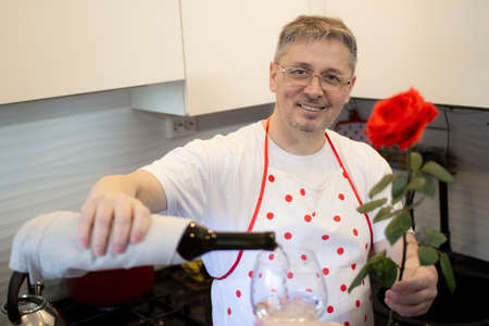 A man in an apron cooks in the kitchen and meets his girlfriend, offering a rose and wine.の写真素材