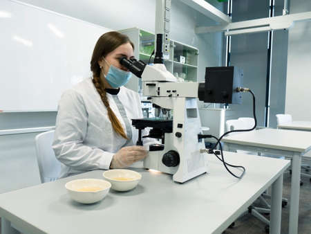 A young woman in a white uniform is working in a laboratory behind a microscope .の写真素材