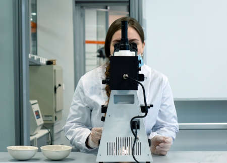 A young woman in a white uniform is working in a laboratory behind a microscope .の写真素材
