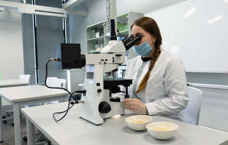 A young woman in a white uniform is working in a laboratory behind a microscope .の写真素材