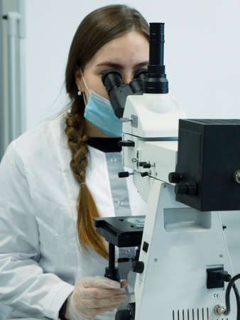 A young woman in a white uniform is working in a laboratory behind a microscope .の写真素材