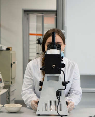 A young woman in a white uniform is working in a laboratory behind a microscope .の写真素材