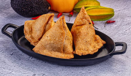 Two crispy fried samosas on a black plate. This popular Indian snack is surrounded by fresh ingredients like bell pepper, star fruit, and chili, creating a vibrant, appetizing food still life.の写真素材