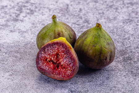 Close-up of green and purple figs on a textured surface. One fig is sliced open, revealing its...