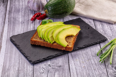 Healthy avocado toast on dark rye bread, served on a black slate board. Rustic scene with fresh...