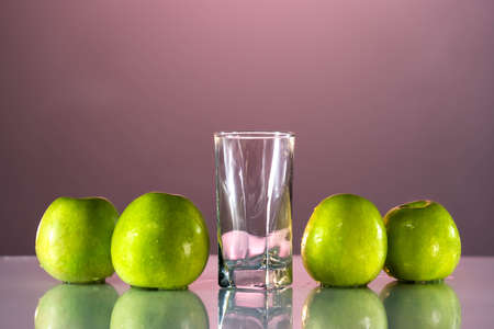Green apples are on the table. next to it is an empty glass for apple juice. Beautiful pink background. Reflected surface of the tableの写真素材