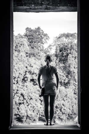 young woman standing in window frame and looking at view through it. Conceptual image for past and futureの写真素材