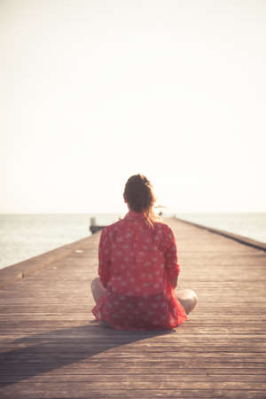 Pensive woman tourist sitting on wooden pier at beach during sunset and looking at infinity view towards horizon and sunset sky during vacation. Back sunlight illuminate woman with copy spaceの写真素材