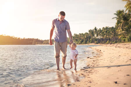 Father and child walking together on the beach coast during sunset with shining sea on background. Father holding child handの写真素材