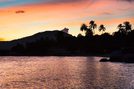 Sunset at tropical beach with palms and hills silhouette and sea ripples on bright orange and blue...