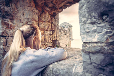 Young woman tourist looking into the distance through ancient stone window in old fortress during sunset in old European city. Concept for people waiting for changes.の写真素材