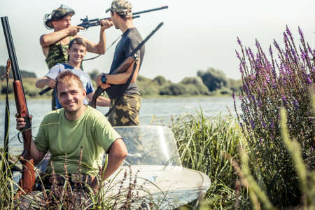 Hunters with guns in boat on river bank during hunting season among reedの写真素材