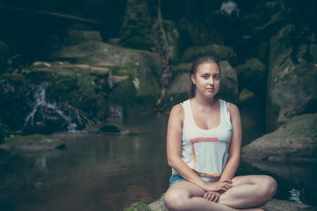 Young woman relaxing in lotus pose on the rocks nearby waterfall with mysterious Mona Lisa smileの写真素材