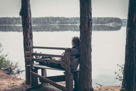 Pensive man on the high edge of river bank sitting on the bench and looking on beautiful scenery with tranquil waterの写真素材