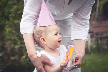 Funny cute baby girl in birthday cap playing with mother in park during birthday celebration.の写真素材