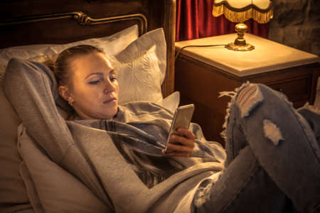 Young  beautiful hipster woman tourist relaxing on bed  in vintage hotel room with mobile phone in...