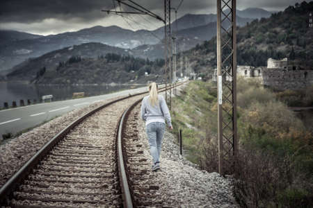 Pensive young woman going away into the distance across railway path in overcast day with dramatic sky  symbolizing parting and despair in relationshipsの写真素材