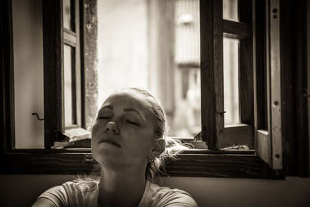pensive young woman with closed eyes lost in thoughts relaxing and dreaming near opened  window in...