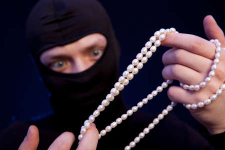 Thief. Man in black mask with a pearl necklace over dark blue backgroundの写真素材