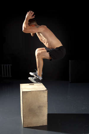 Man jumping on fit box in gym. Male with naked torso exercises jump on the dark backgroundの写真素材