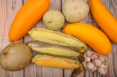 An unshelled ear of corn, young garlic in a wicker basket, marble pumpkins and yellow zucchini on...