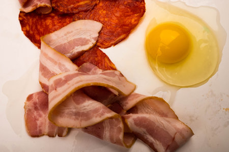 A broken chicken egg without a shell, slices of sausage and bacon and tomatoes on a white...