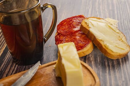 Fresh wheat toast with butter, sausage and cheese, a wooden butter dish with a piece of butter and a mug of tea on a wooden background. Close upの写真素材