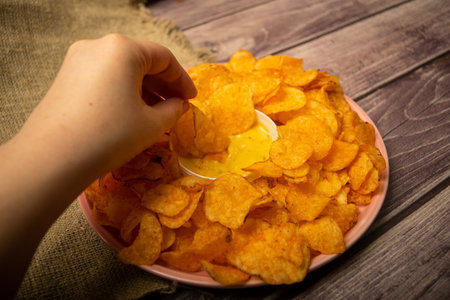 The girl takes a chip from a round dish with potato chips and a saucepan with cheese sauce in the...