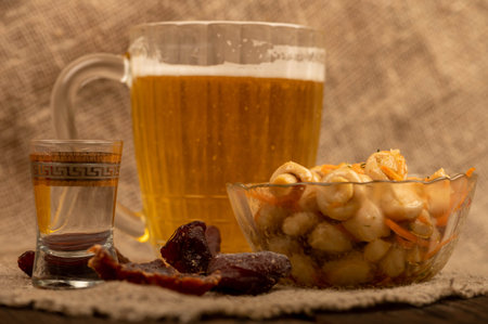 Pickled mushrooms in a glass dish, a glass of vodka and a mug of beer on a table covered with a...