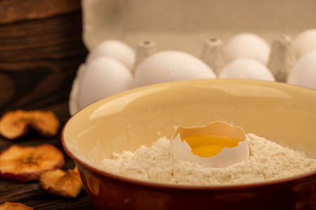A broken white chicken egg in a bowl with wheat flour, dried apple chips and several whole eggs in a tray made of white cardboard, close-up selective focusの写真素材